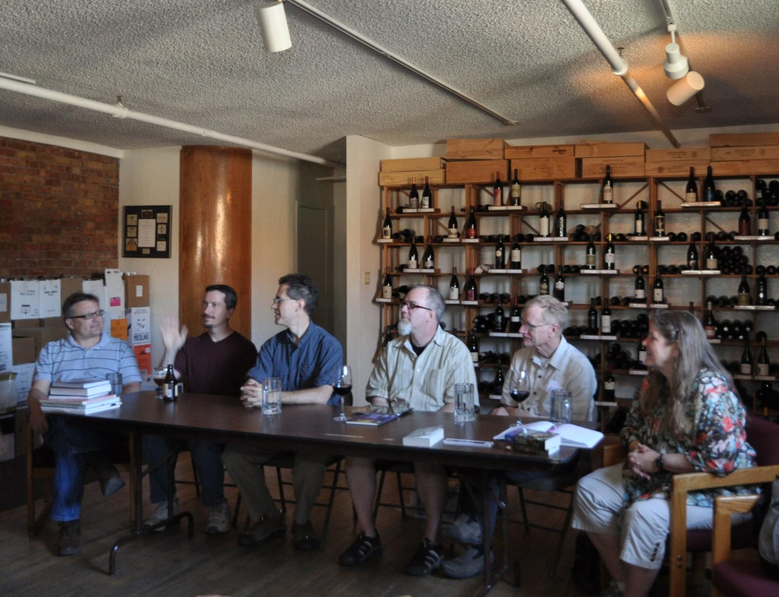 Five men and one woman in casual clothing sit behind a long folding table in a room with a shelves full of wine bottles behind them.