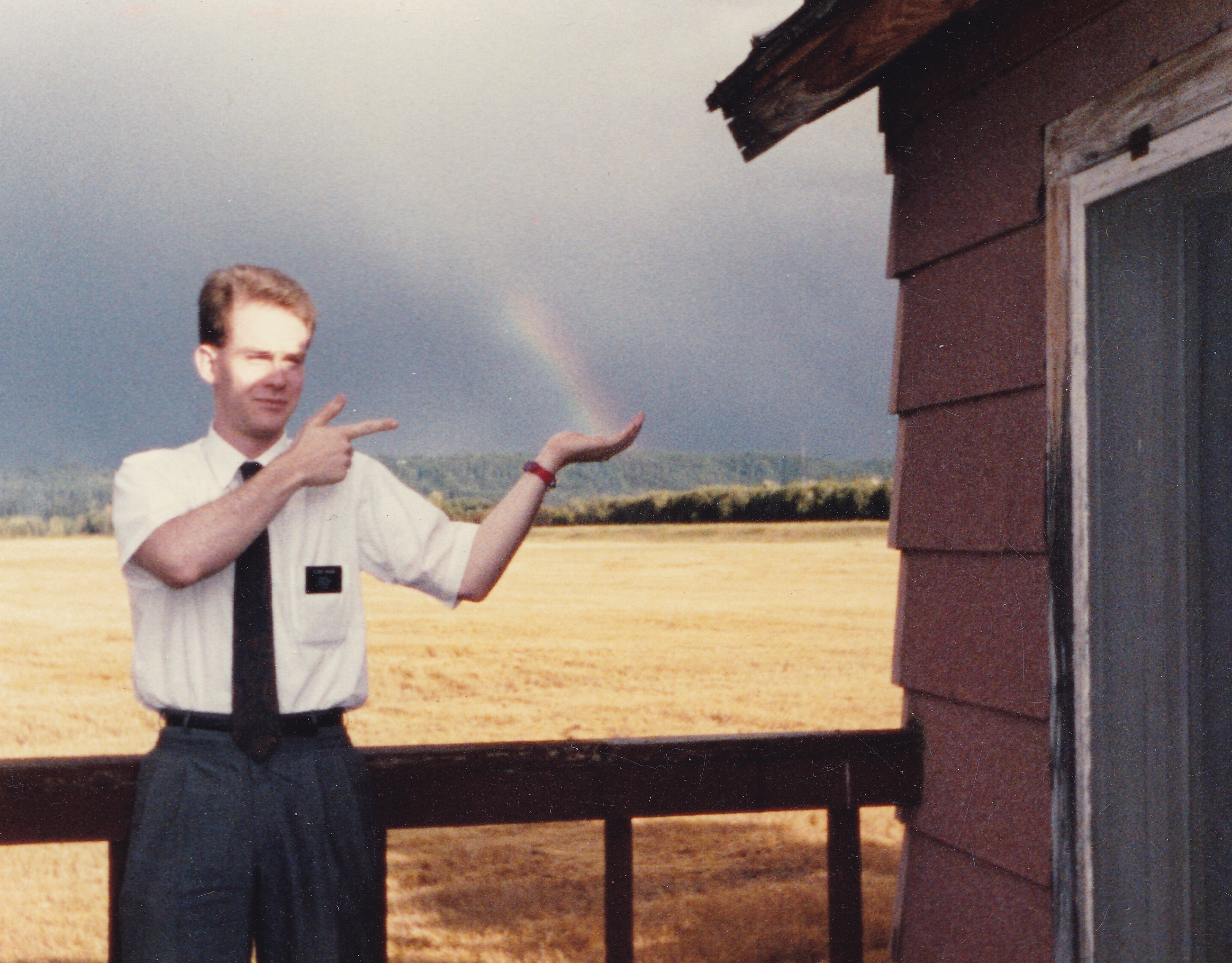 A Mormon missionary in white shirt and tie stands on the deck of a small house in front of a wheat field, appearing to hold a rainbow in his hand.
