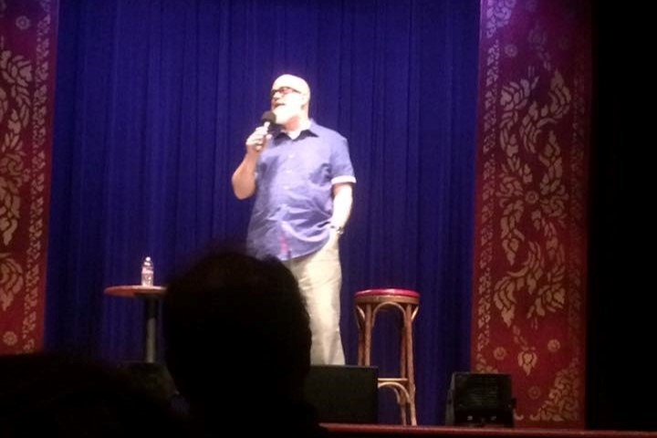 A bearded man stands on stage near a stool in front of a blue curtain in a classic-style theater, with the silhoutted heads of audience members in the foreground.