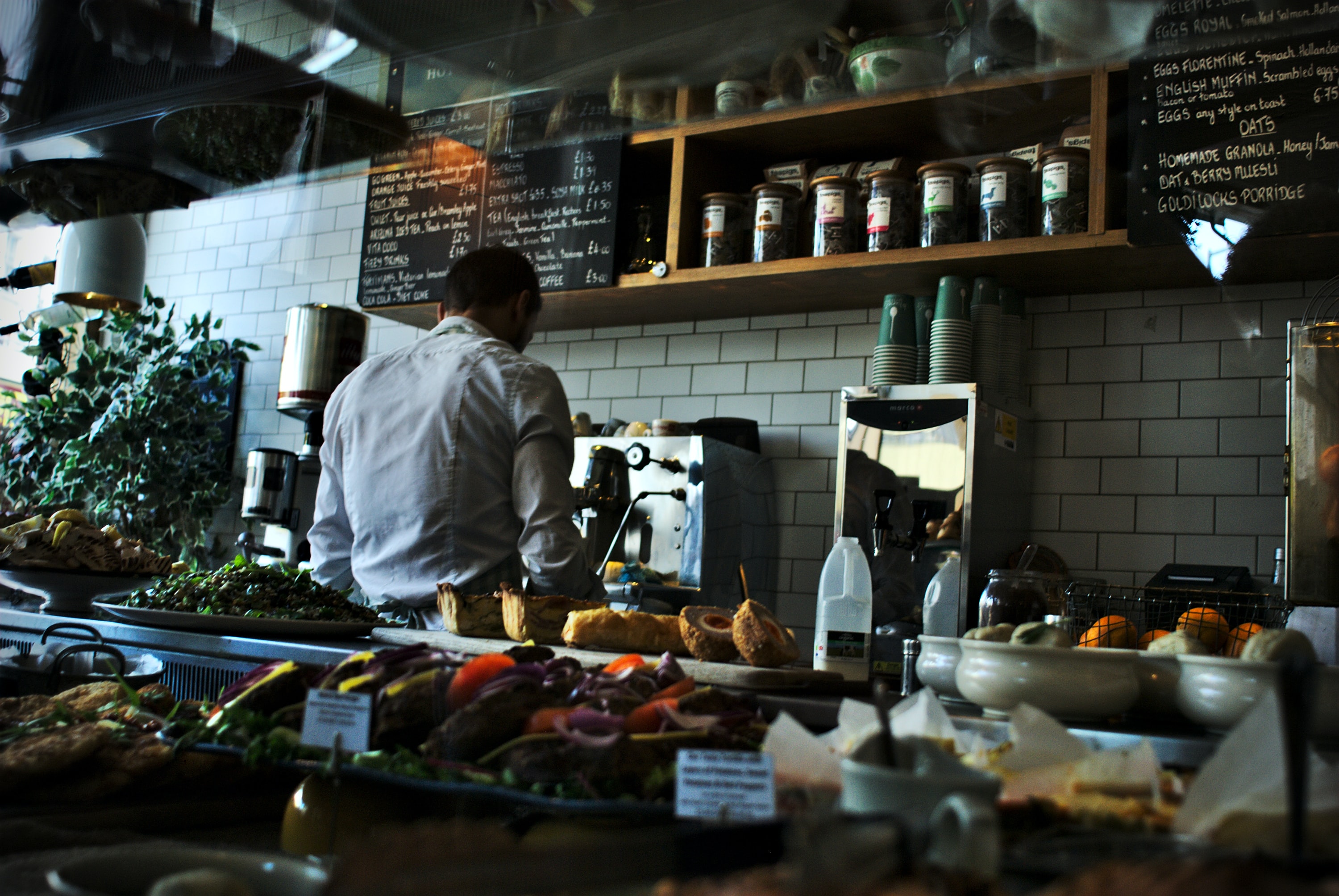Behind a counter crowded with food displays, a chef's back is turned as they prepare food in a fancy delicatessen.