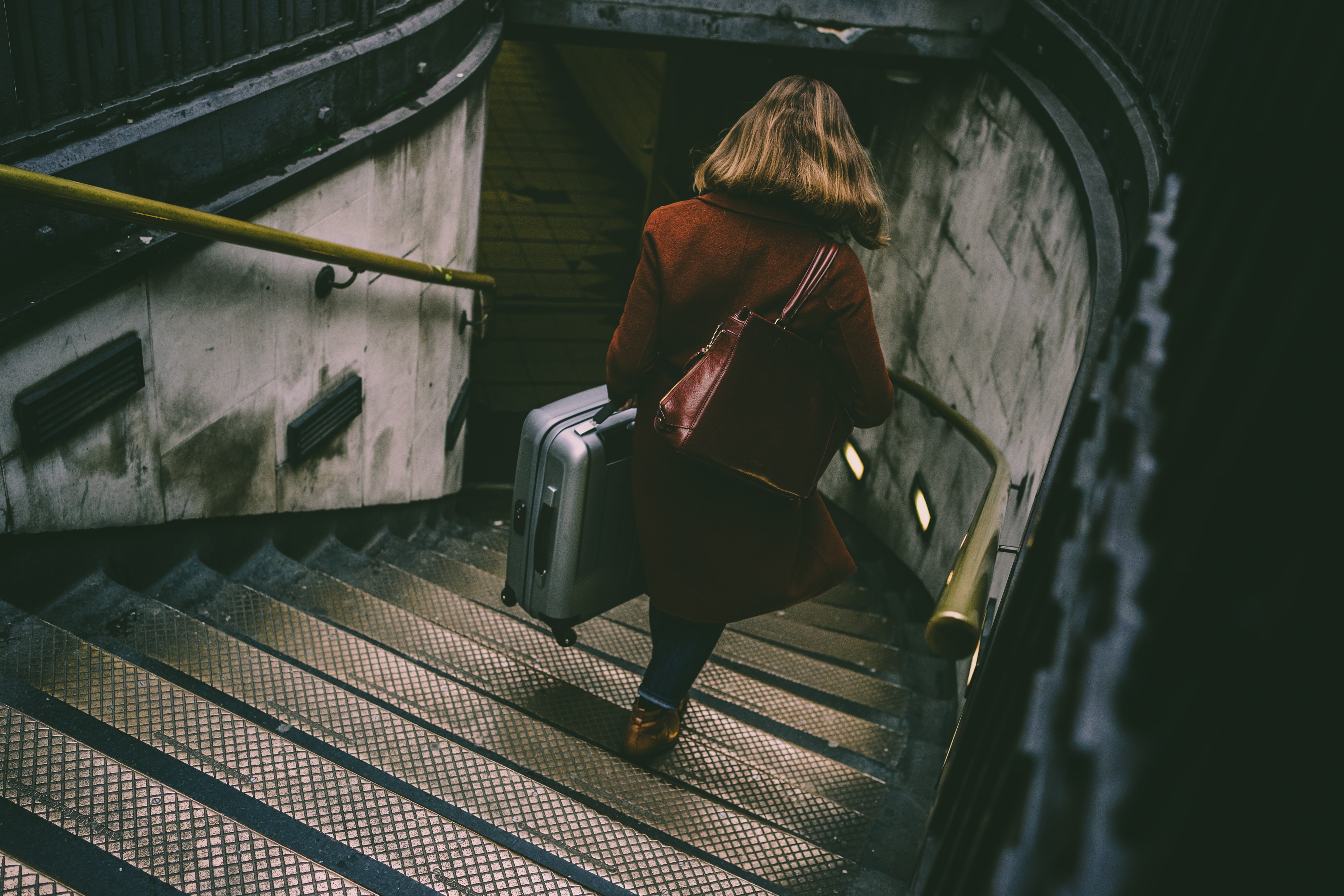 A young woman, viewed from above and behind, dressed for chilly weather, carries a suitcase down a gray and discolored staircase into an urban train station.
