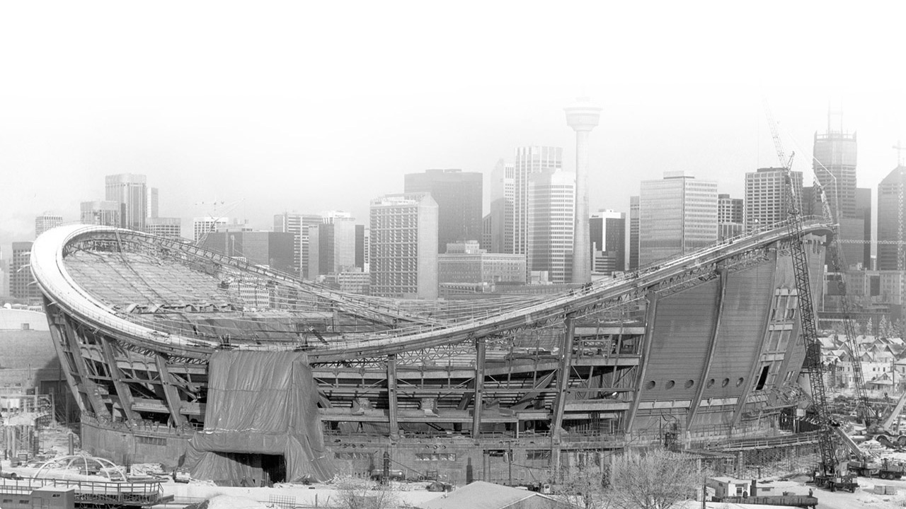 Black-and-white photograph of Calgary's Saddledome, a saddle-shaped sports arena, in mid-construction, with the city skyline in the background.